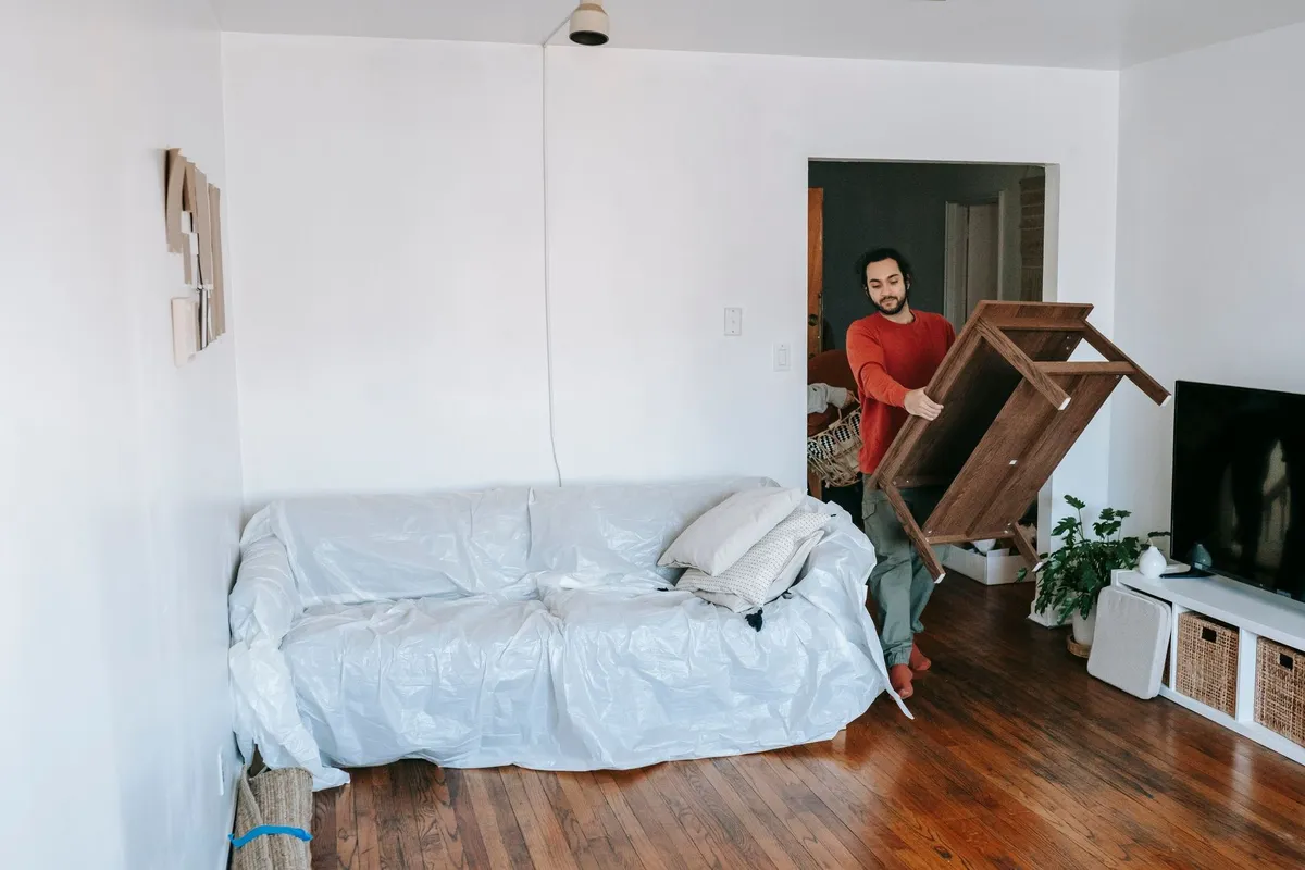 A man lifts a wooden table in a bright living room with a wrapped sofa, illustrating furniture removal.